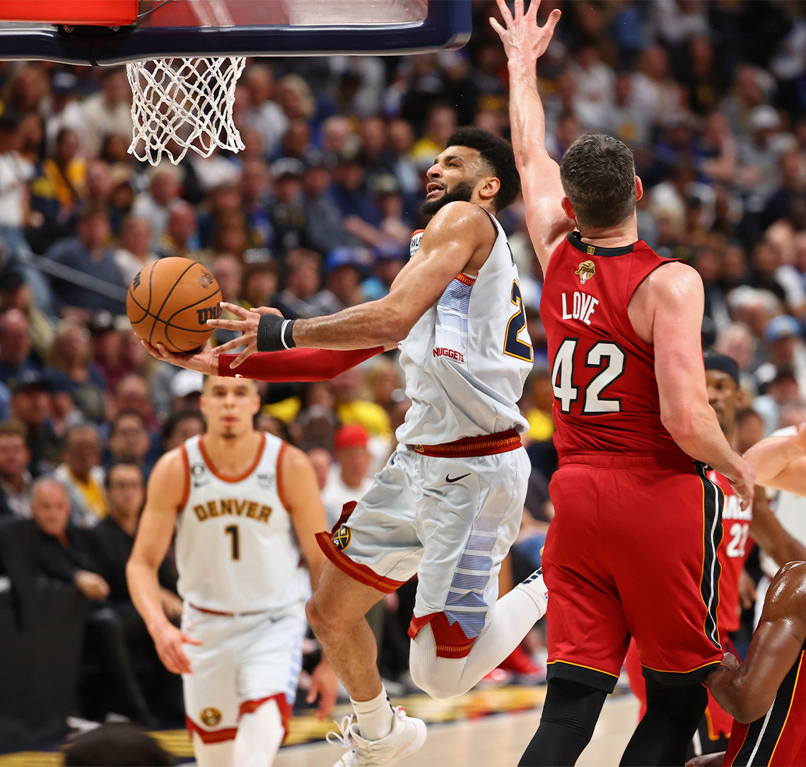 Jamal Murray jumps to throw the ball into the net. An opposing player reaches up to defend, while a team mate looks on.