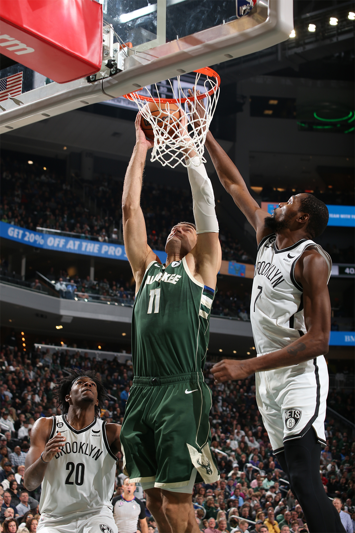 Brook Lopez jumps to dunk the basketball into the net. Another player observes offside, and another jumps up to defend.