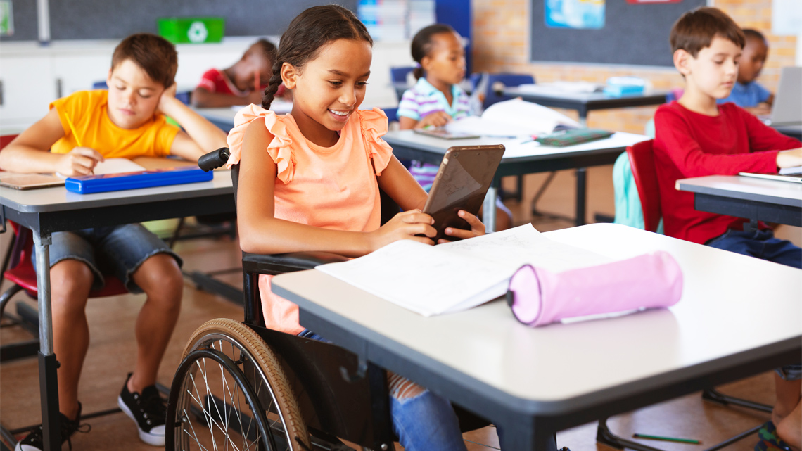 Children seated at their desk doing various activities, like reading, drawing, or using their devices, like a tablet or a laptop.