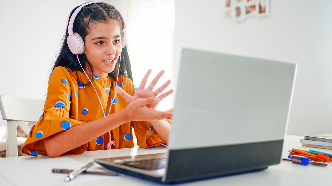 Learner wears headphones that are connect to a laptop to communicate with others. Learner is participating in an online class from home.