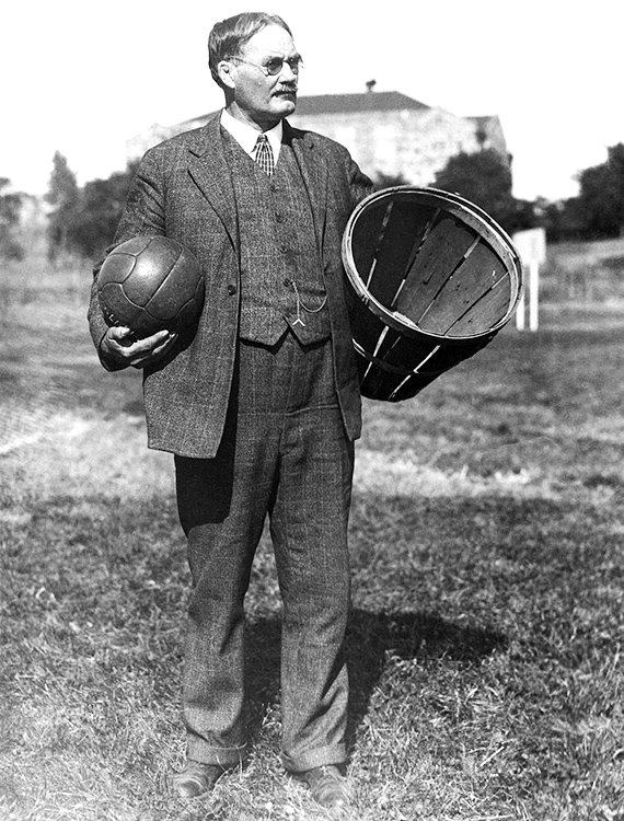 James Naismith standing and holding a basketball and a large peach basket.