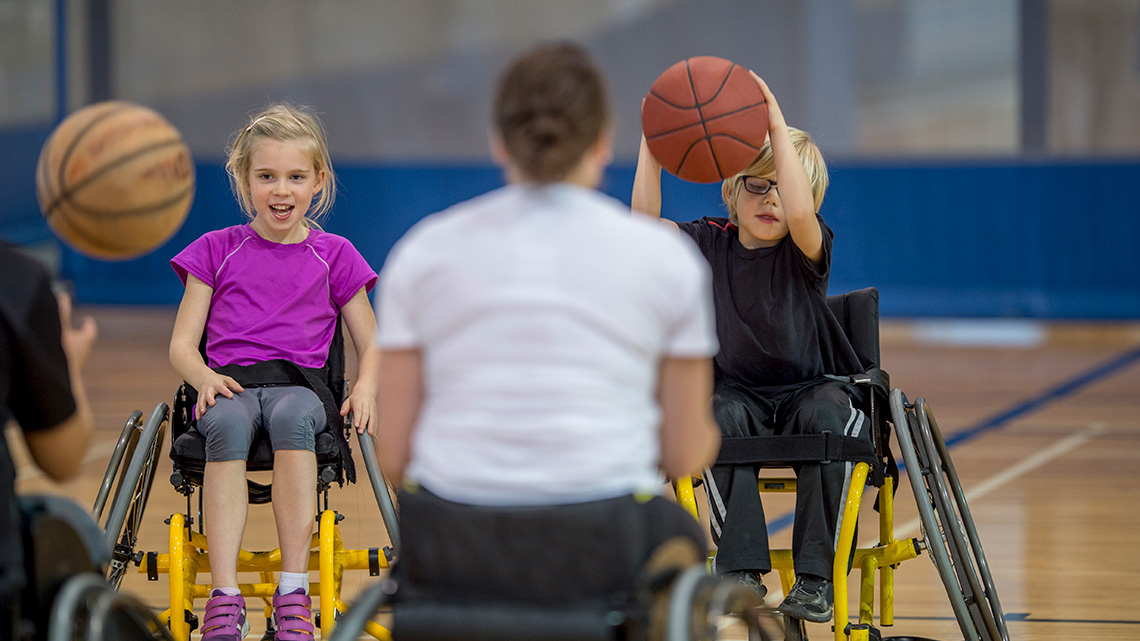 Differently abled children seated in wheelchairs, while dribbling basketballs in a basketball court.