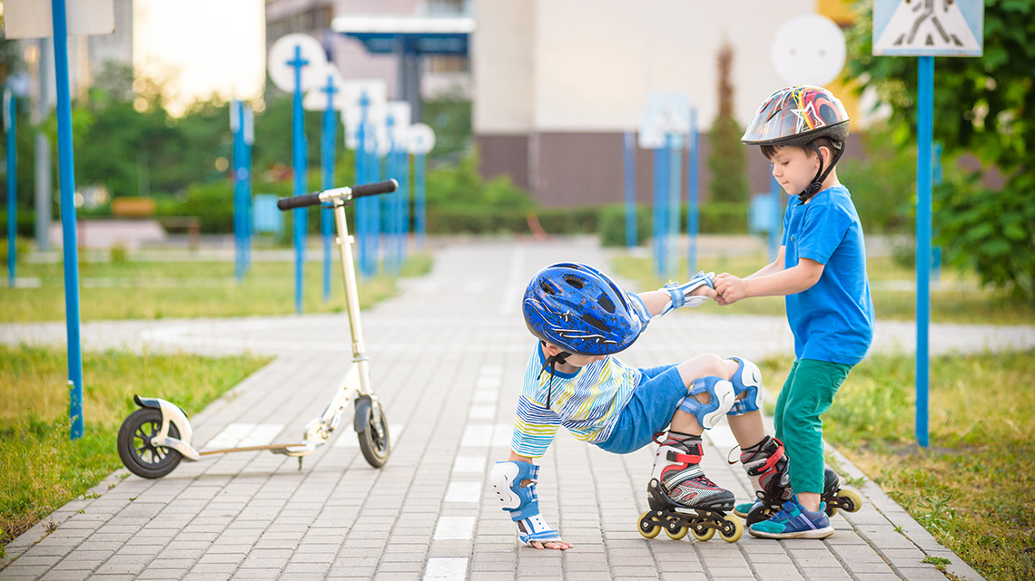 Two children roller skating and scootering. One child helps another but pulling them up from the ground.