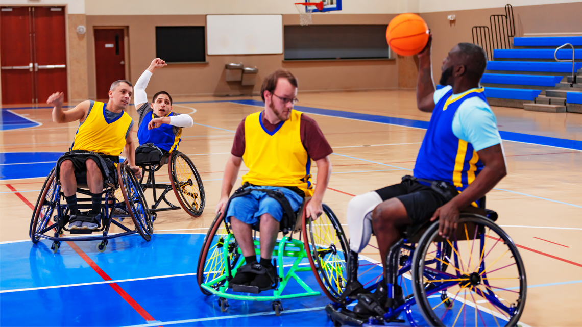 Wheelchair basketball teams play basketball in a court. Wheelchair has large, slanted wheels, small rear and front wheels, and no armrests.