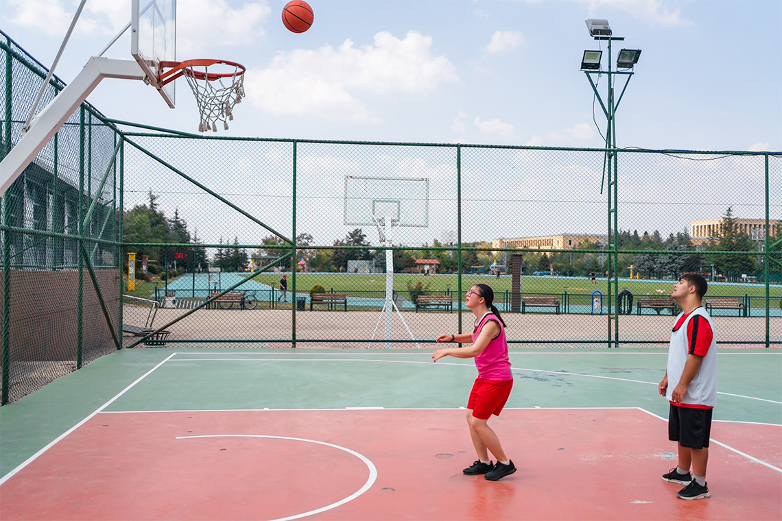 Two individuals playing basketball on a basketball court with the basketball mid-air towards the hoop.