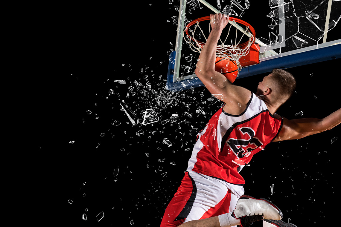 Basketball player dunking basketball while the backboard shatters. Player holds onto the rim.