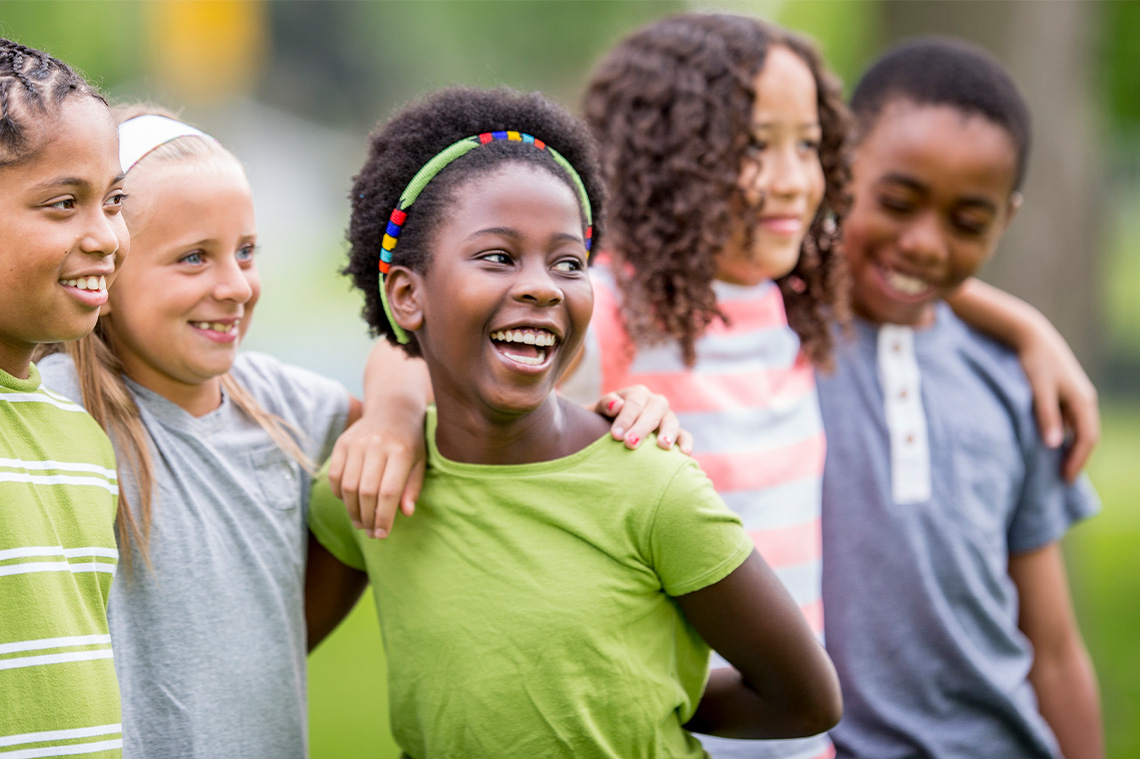 Children laughing and smiling as they stand together with arms over each other’s shoulders.