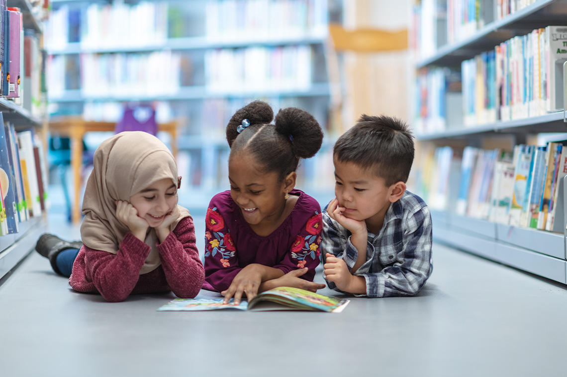 Children smiling and thinking as they read a book together in a library.
