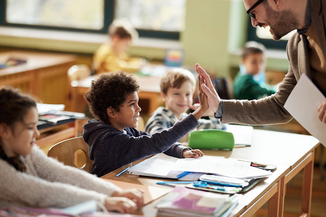 Teacher giving a student a high-five in the classroom. Both teacher and students are smiling.