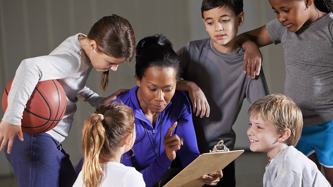 A group of young basketball players surround their coach. Their coach reads off a clipboard while the players listen.