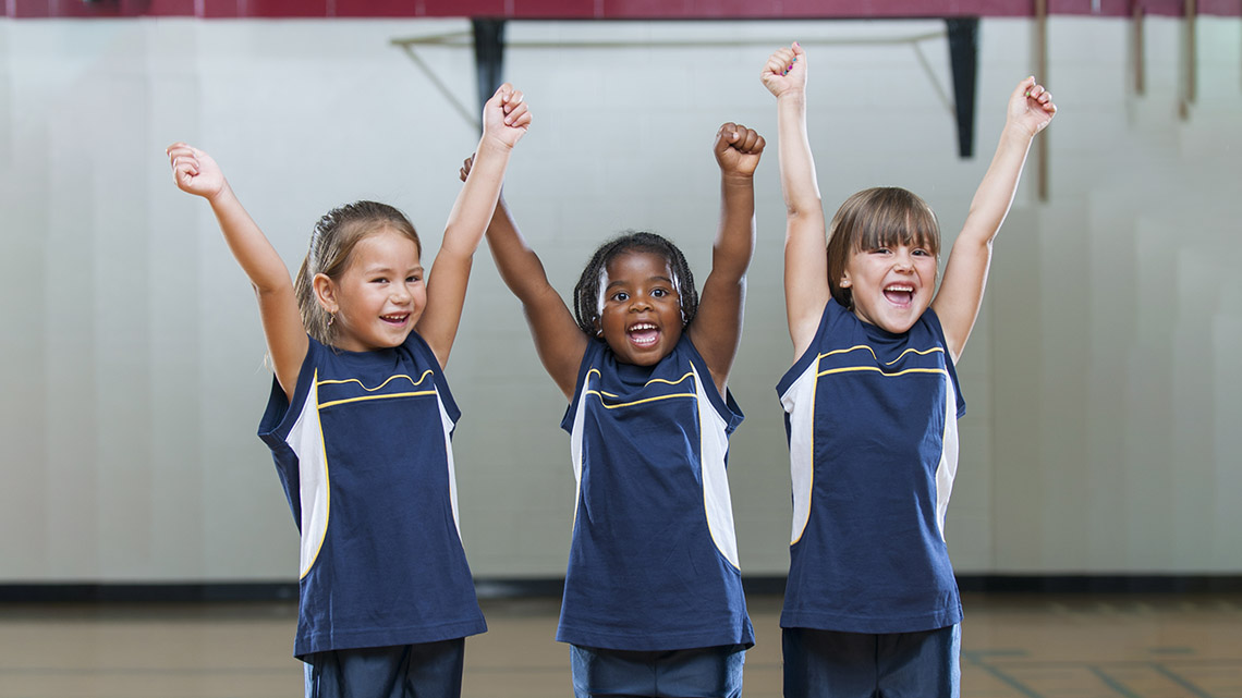 Three young basketball players smile as they stand with their hands raised on a basketball court.