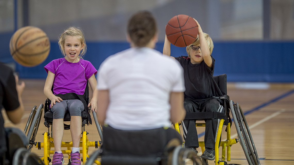 On a basketball court, two young basketball players in wheelchairs pass basketballs to their coaches. They throw the ball using two hands.
