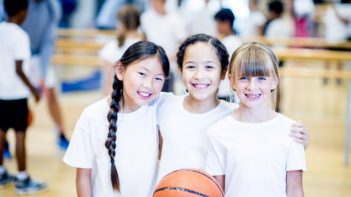 Three young students stand together and smile. One holds a basketball. In the background, other players listen to their coach on the court.