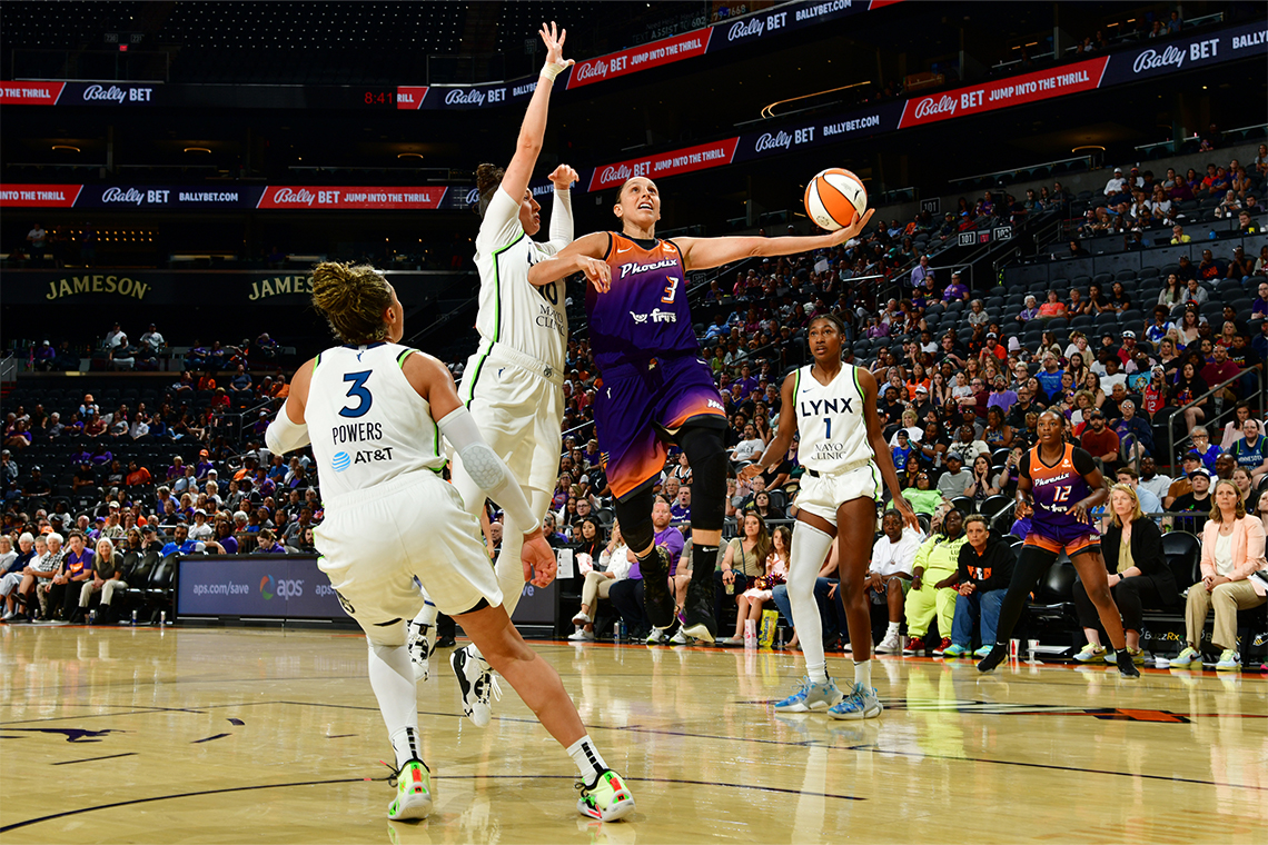 Diana Taurasi jumps up to drive the ball into the net. A player jumps with stretched arms, while another runs over. A teammate looks on.