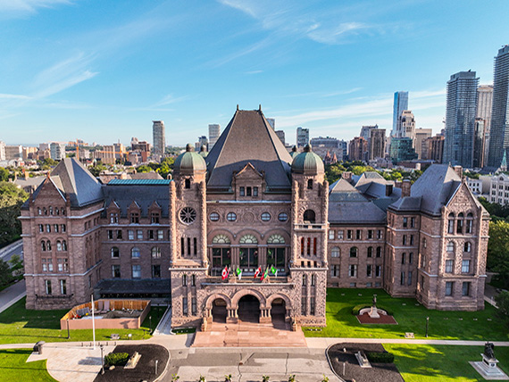A large stone building in a city park features round arches and a central tower. The main entrance is framed by large columns and archways
